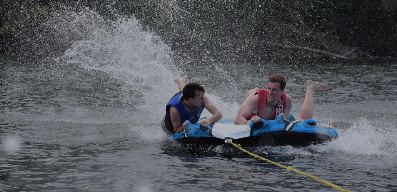 Image: Two camp Trulaske students on an inter tube being pulled on a lake.