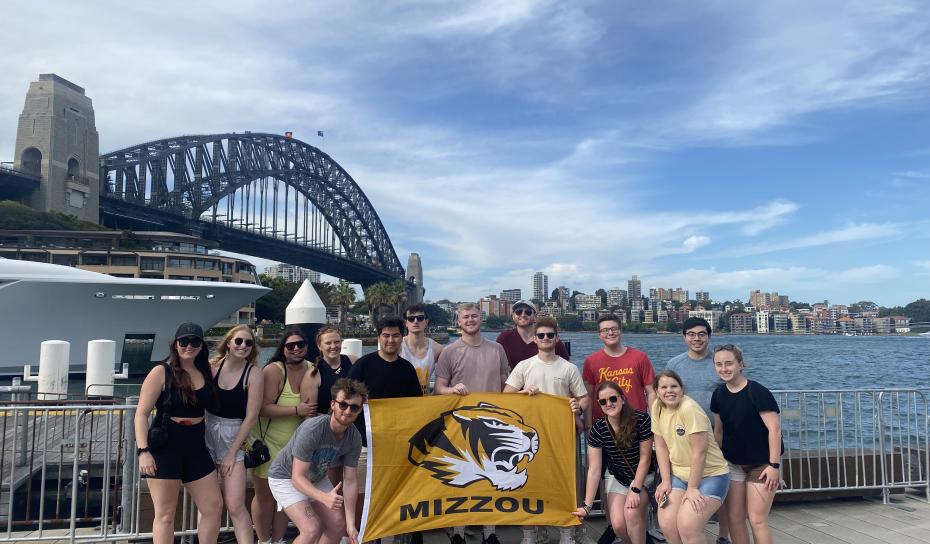 Group on students holding MU flag in front of Sydney Harbor Bridge