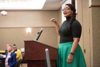 Image: An African American woman standing at a podium speaking to a seated crowd.
