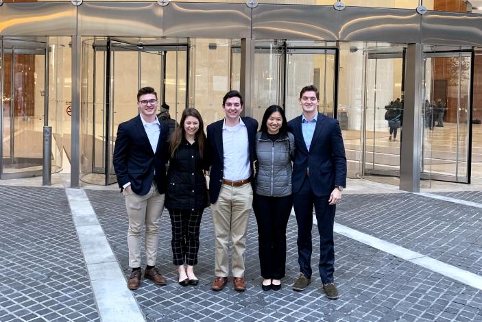 Image: A group of students standing a front of a building's glass entrance. The sign "Deloitte" is visible inside the building.